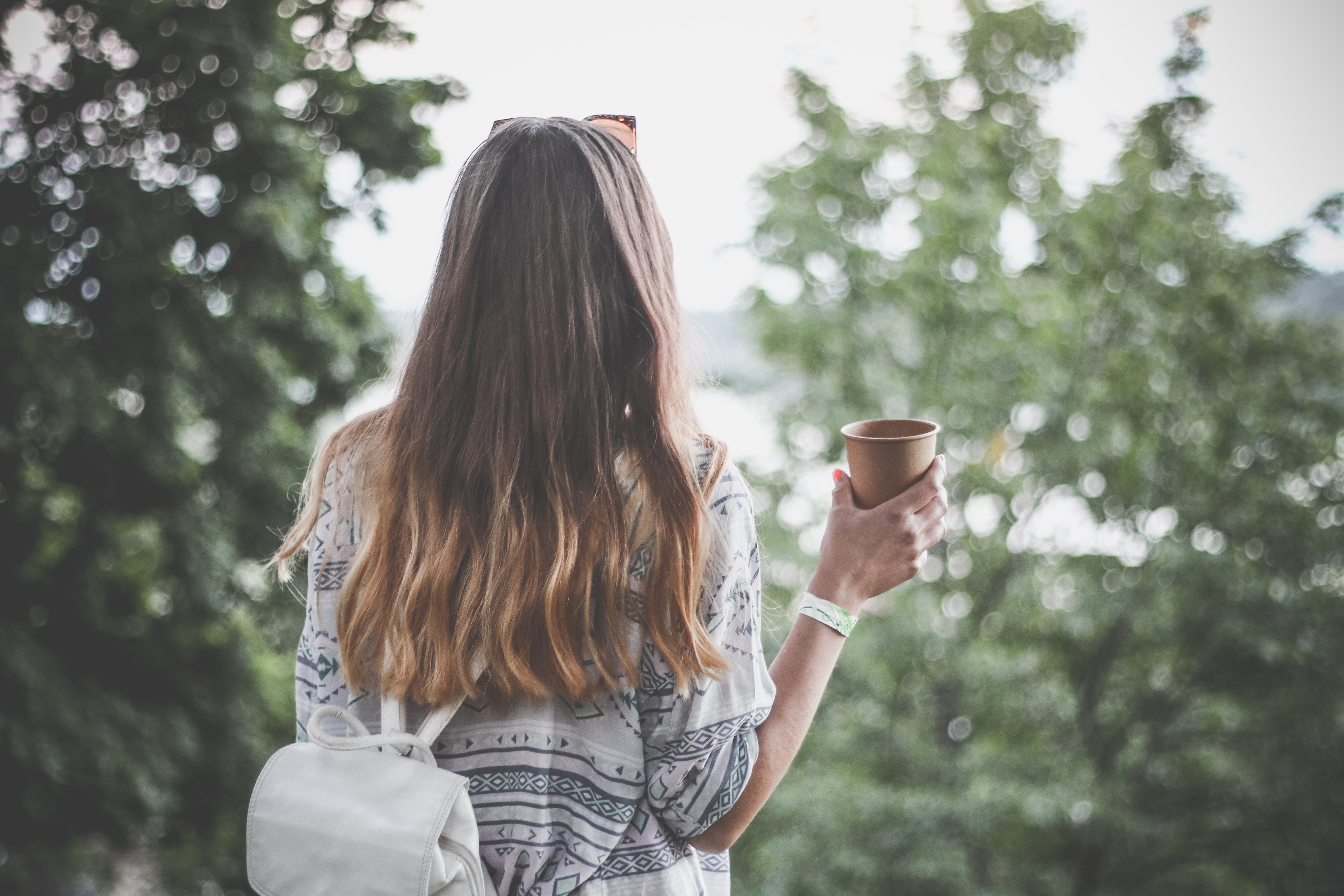 A woman standing, her back facing the camera, holding a cup looking out at trees and nature. Woman represents the use of christian mindfulness.